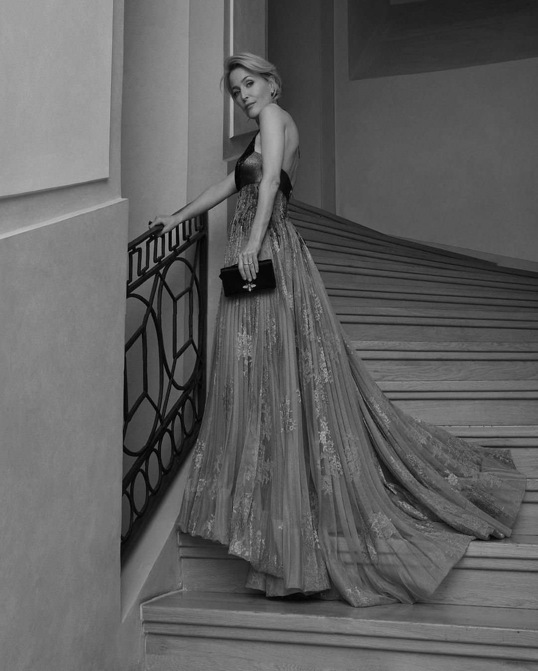 Gillian Anderson wearing a flowing evening gown on a grand staircase, holding a small clutch by Andreas Ortner