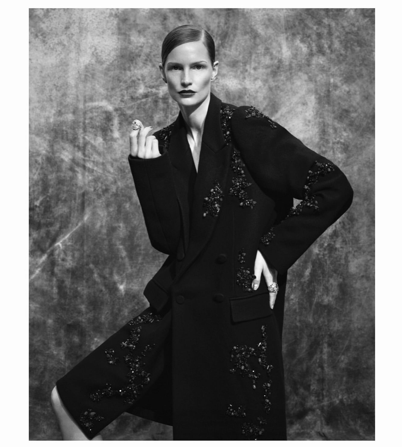 Model in dark embellished coat with beaded appliques against a textured grey studio background