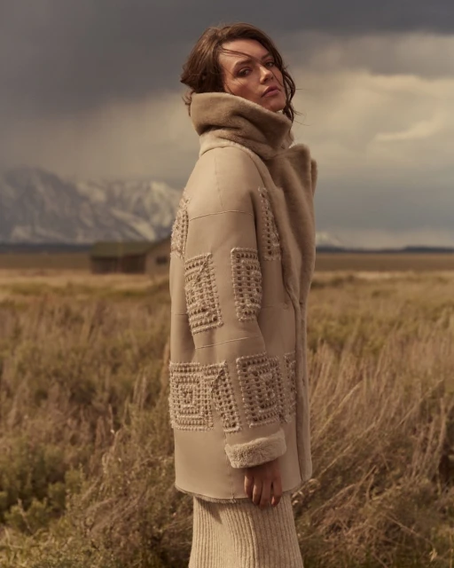 Woman in embroidered coat posing in golden field with mountains, cloudy sky. Fashion photography in an outdoor setting, medium shot.