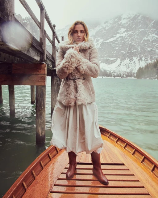 A woman in a fur-lined coat stands on a wooden boat in a tranquil lake, surrounded by snow-capped mountains and a wooden pier.