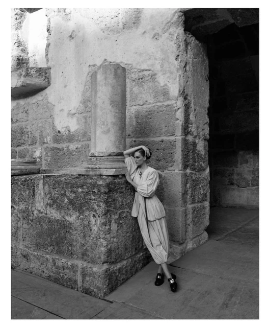 Black-and-white fashion shot of a woman posing in a flowing outfit against an old stone wall and pillar. Editorial fashion photography.