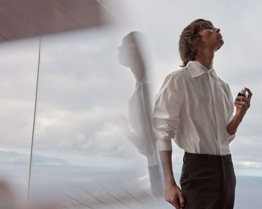 Man in white shirt sprays cologne near a window overlooking the ocean, creating a reflection. Fashion and beauty product advertisement.