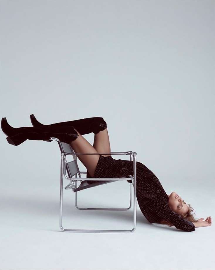 Fashion shot of a female model wearing black heeled boots and a dark minidress reclining in a chrome chair against a white backdrop.