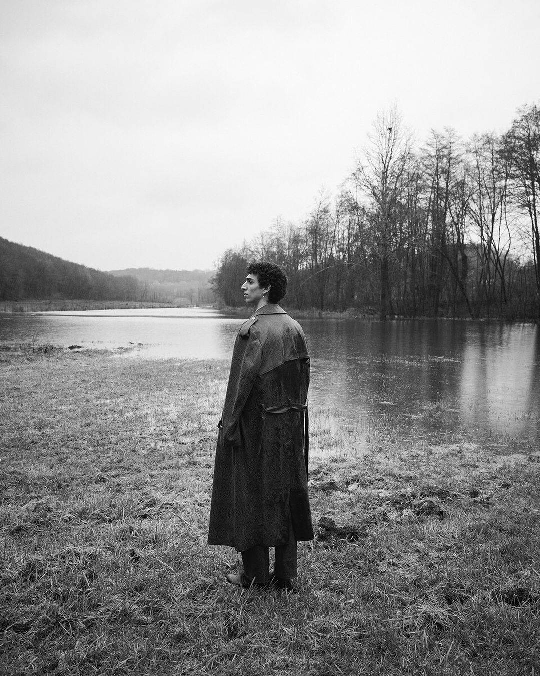 Monochrome outdoor portrait of a lanky adult standing in a trench coat in a grassy, partially flooded rural landscape with a water backdrop.