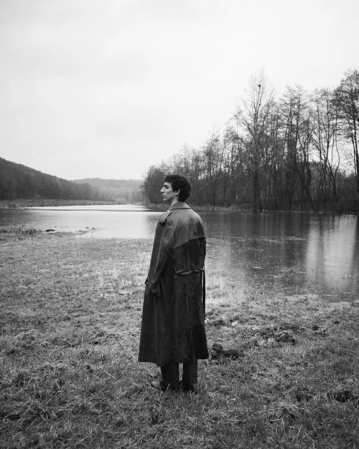 Monochrome outdoor portrait of a lanky adult standing in a trench coat in a grassy, partially flooded rural landscape with a water backdrop.
