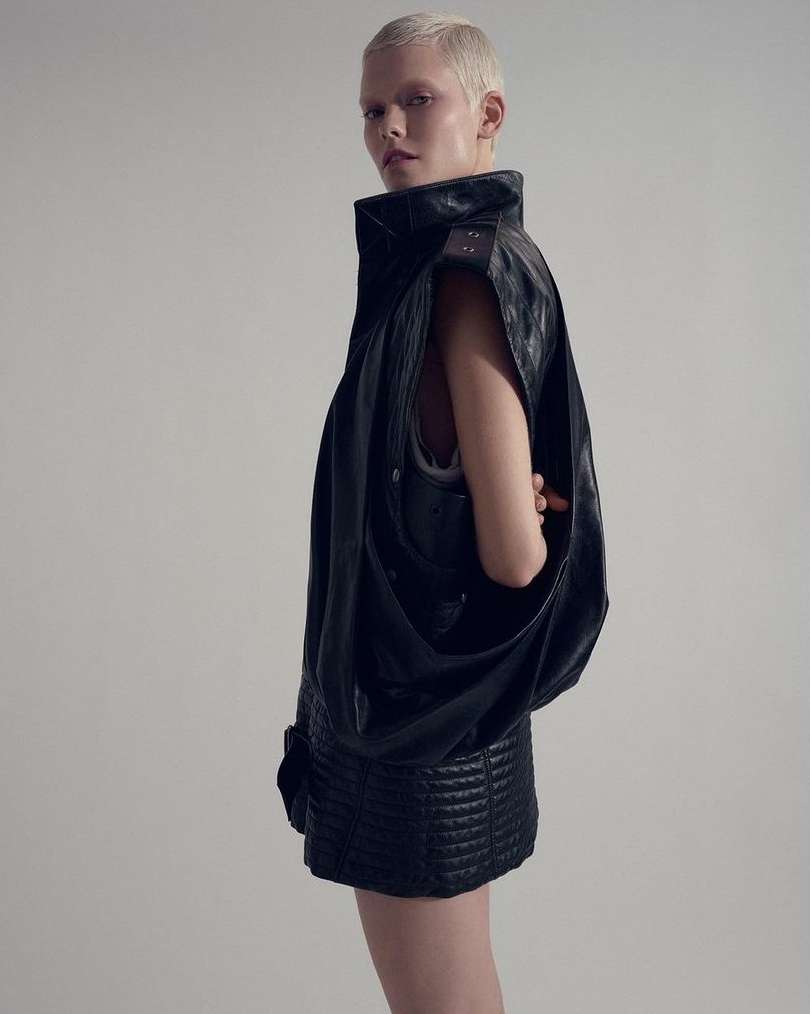 Blonde woman with short hair poses in a black leather sleeveless jacket and a short skirt against a white background in a studio shot.