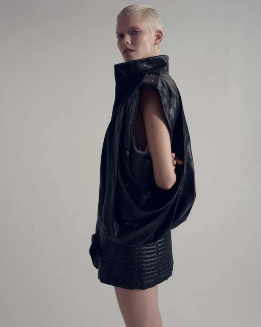 Blonde woman with short hair poses in a black leather sleeveless jacket and a short skirt against a white background in a studio shot.