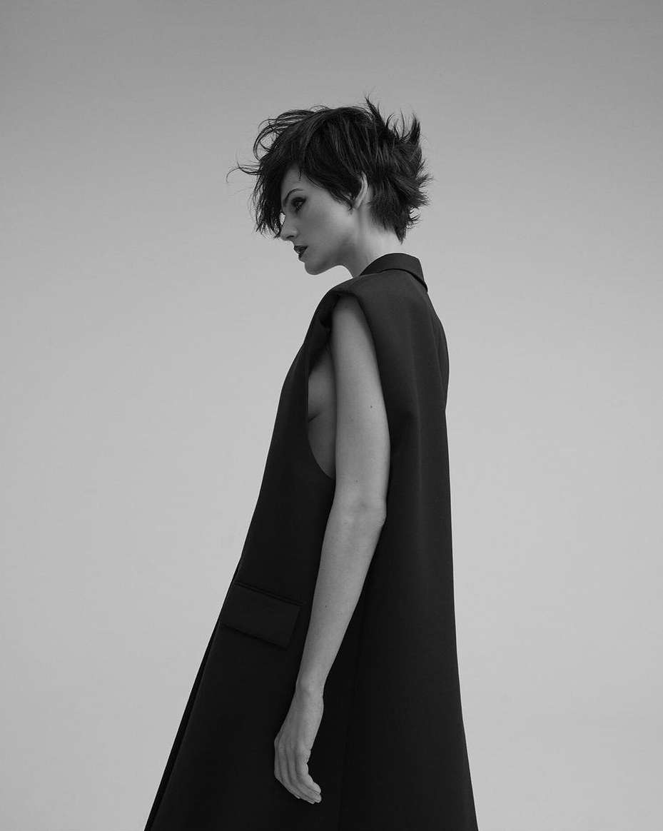 Monochrome portrait of a woman with short hair wearing a long black vest, standing in a light grey photo studio looking to the left.