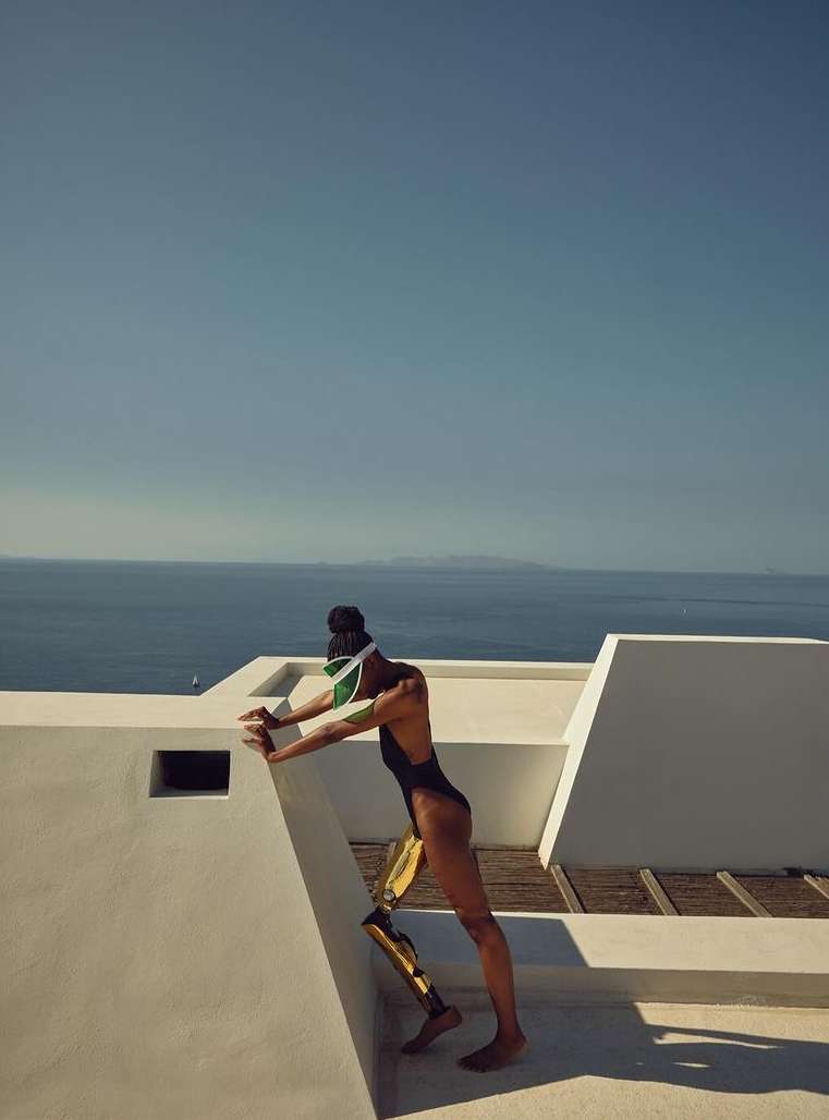 Athletic woman with prosthetic leg in swimsuit stretches against white wall overlooking the ocean, sunny sky, and outdoor setting.