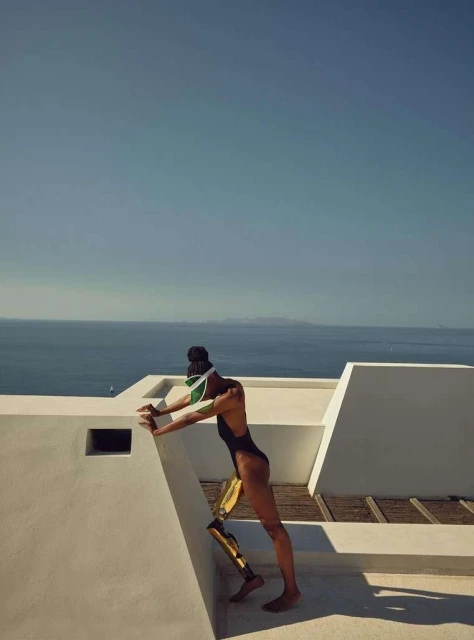 Athletic woman with prosthetic leg in swimsuit stretches against white wall overlooking the ocean, sunny sky, and outdoor setting.