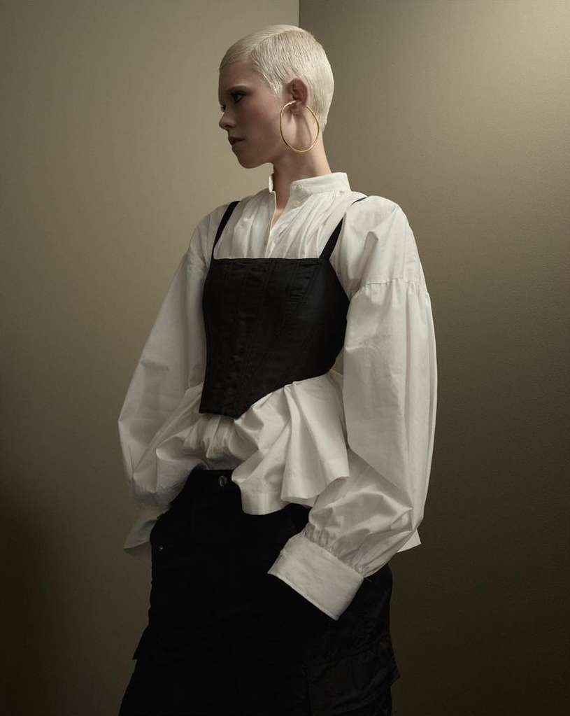 Blonde-haired person wearing a white blouse and black top in a studio setting. Fashionable clothing with subtle shadows.