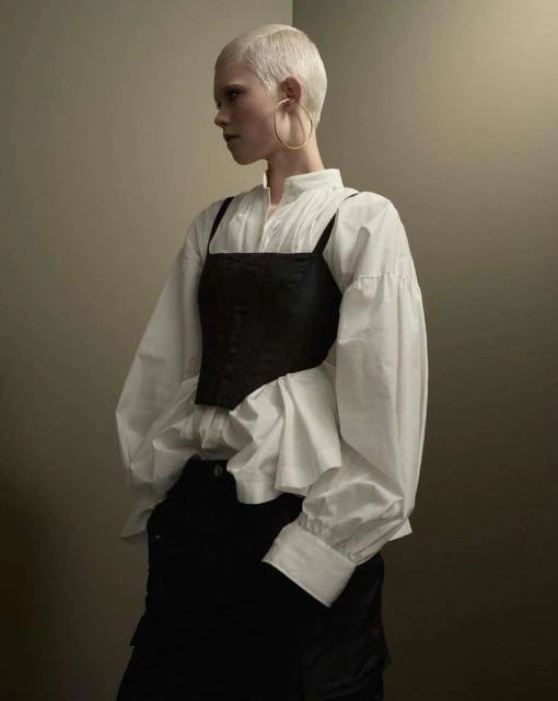 Blonde-haired person wearing a white blouse and black top in a studio setting. Fashionable clothing with subtle shadows.