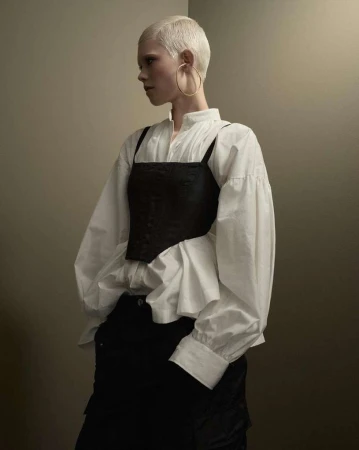 Blonde-haired person wearing a white blouse and black top in a studio setting. Fashionable clothing with subtle shadows.