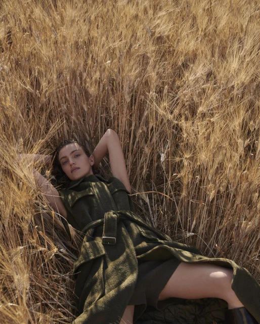 Woman in green dress lying in golden wheat field, looking at the camera. Outdoor fashion portrait in a rural landscape setting.