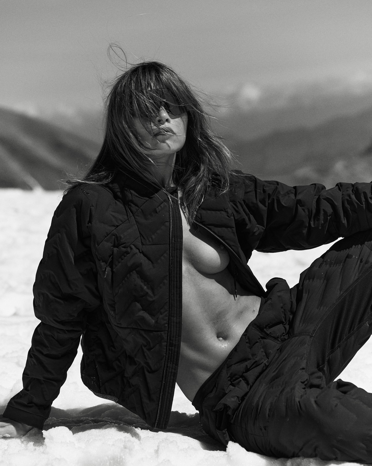 Monochrome portrait of a reclining adult woman in a black quilted jacket and pants in a snowy mountain landscape on a sunny day.