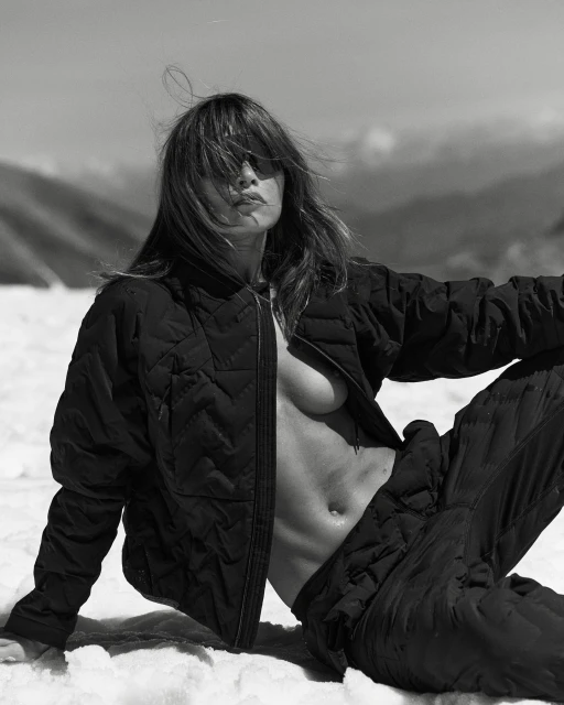 Monochrome portrait of a reclining adult woman in a black quilted jacket and pants in a snowy mountain landscape on a sunny day.