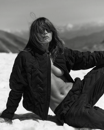 Monochrome portrait of a reclining adult woman in a black quilted jacket and pants in a snowy mountain landscape on a sunny day.