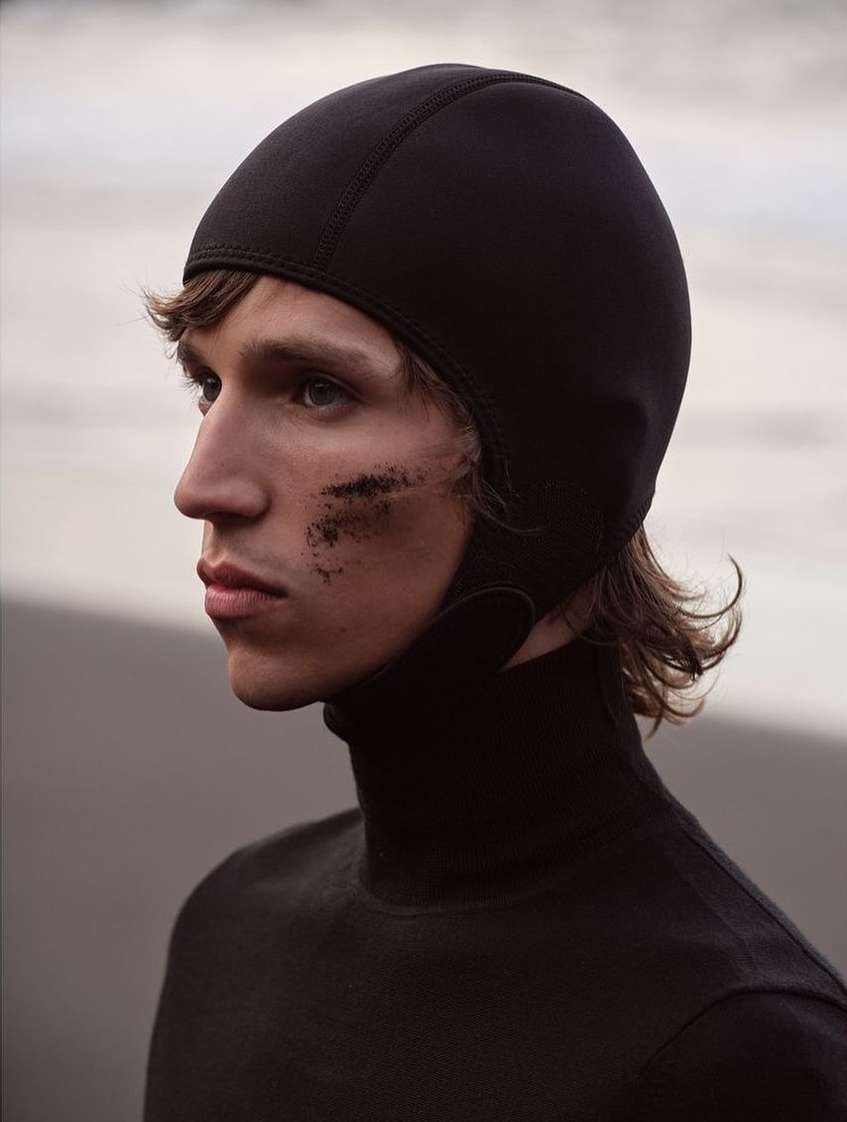 Close-up profile of a male model wearing a black hood and turtleneck with dark smudges on his cheek against an out-of-focus beach background.