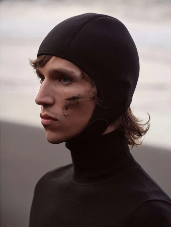 Close-up profile of a male model wearing a black hood and turtleneck with dark smudges on his cheek against an out-of-focus beach background.