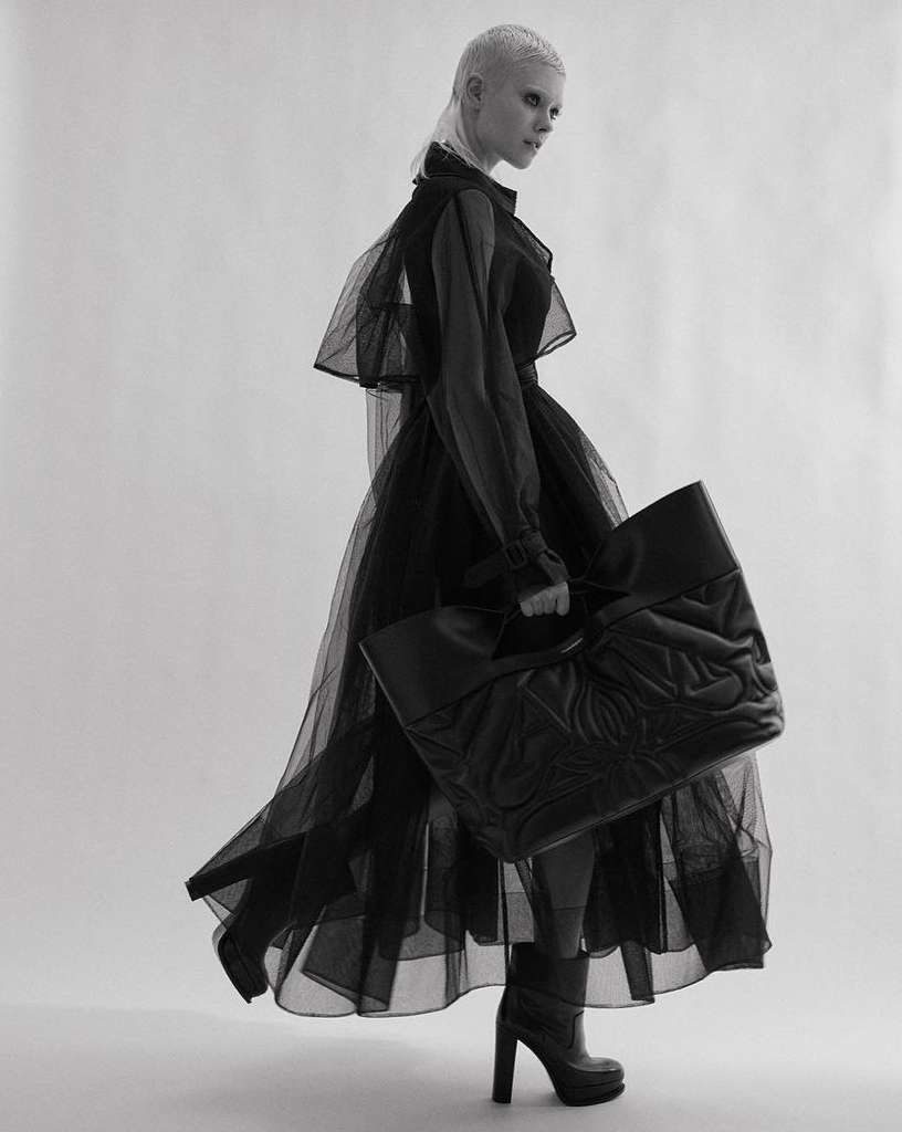A woman in a sheer black dress poses with a large black tote bag in a studio, creating a high-fashion editorial shot against a white background.
