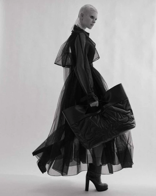 A woman in a sheer black dress poses with a large black tote bag in a studio, creating a high-fashion editorial shot against a white background.