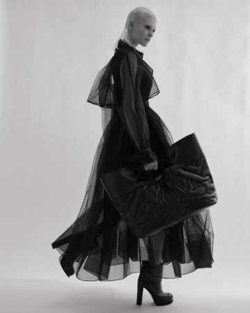 A woman in a sheer black dress poses with a large black tote bag in a studio, creating a high-fashion editorial shot against a white background.