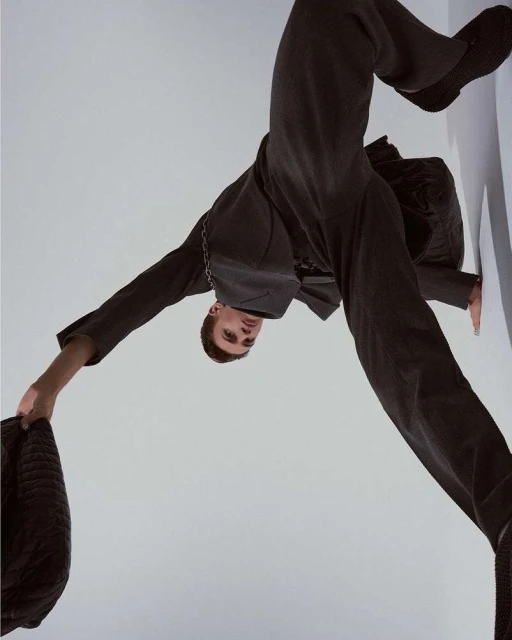Upside-down portrait of a young adult man in a dark suit holding a jacket against a light grey wall with soft studio lighting.