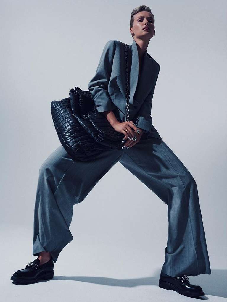 Woman in a grey suit and black loafers poses with a quilted leather bag in a studio environment, showcasing fashion and editorial style.