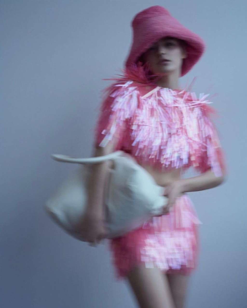 Woman in pink fringe outfit and matching hat poses with a white purse against a grey background in a fashion editorial style image.