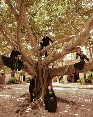 Group of five people posed in and around a large tree in a courtyard with a building in the background, all dressed in black clothing.