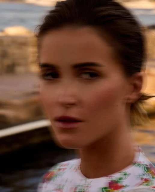 Woman gazes at camera outdoors with blurred motion. She has brown hair, fair skin, and wears a floral-patterned dress.