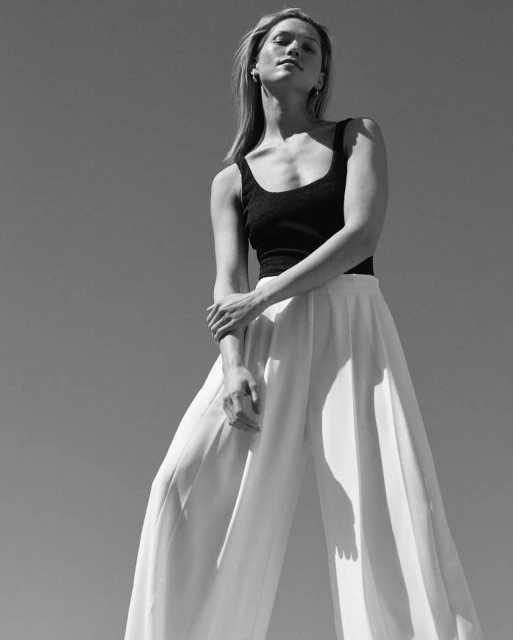 Monochrome portrait of a young adult woman in a black top and white wide-leg pants against a bright sky background, low angle.