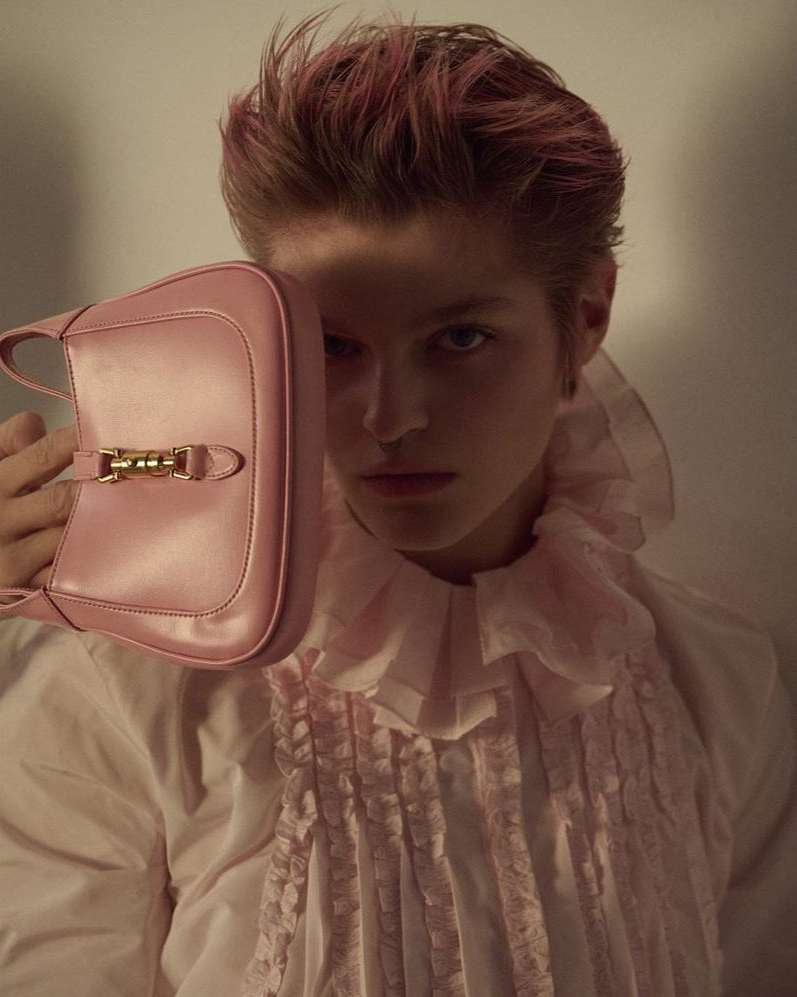 Person posing indoors with a pink purse and pink-tinted hair, wearing a white ruffled shirt, with a light neutral background.