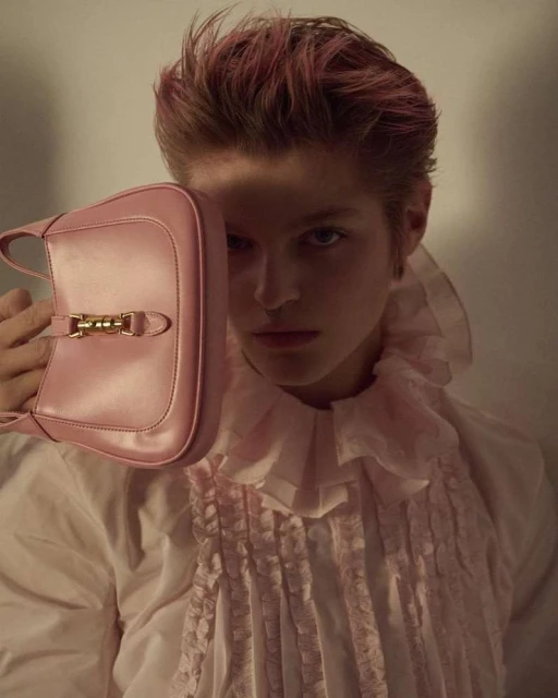 Person posing indoors with a pink purse and pink-tinted hair, wearing a white ruffled shirt, with a light neutral background.