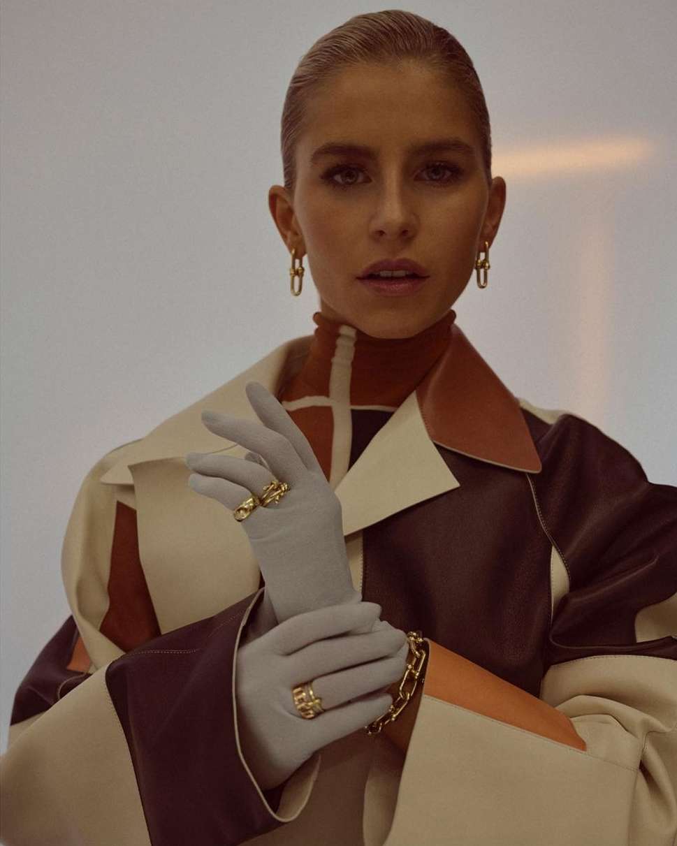 Fashionable woman in studio wearing statement gold rings and jewelry, elegant gloves and designer coat in front of a light neutral background.