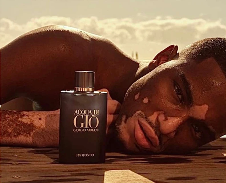 Close-up shot of Armani Acqua di Gio fragrance bottle with dark-skinned man lying on a wooden surface under a bright sky.