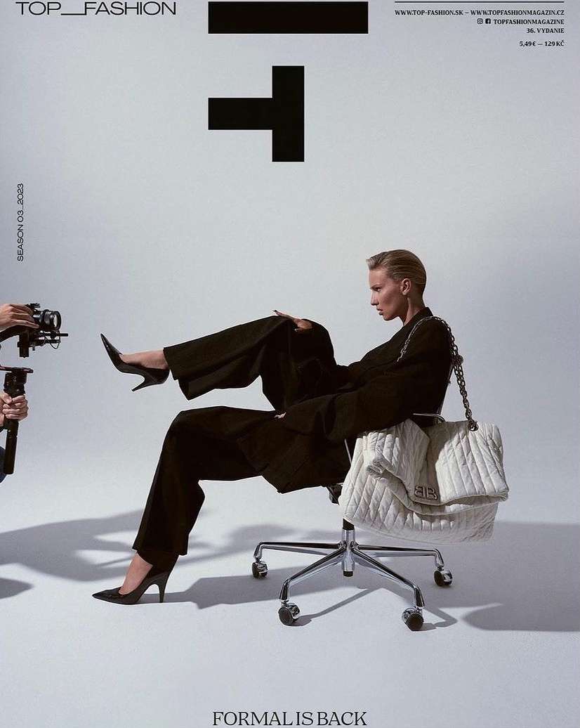 Fashion editorial featuring a woman in a black suit and heels, posed on a rolling chair with a large white quilted bag, in a white photo studio.