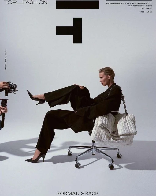 Fashion editorial featuring a woman in a black suit and heels, posed on a rolling chair with a large white quilted bag, in a white photo studio.