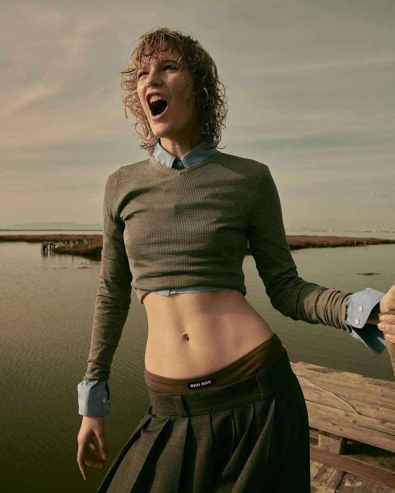 Fashion image of a young adult female model with curly hair, wearing a crop top and pleated skirt, posing near a body of water.