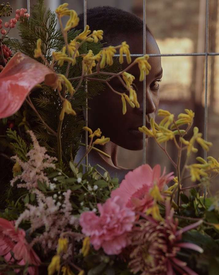 Portrait of a Black woman framed behind a wire fence and lush arrangement of pink and yellow flowers by Andreas Ortner