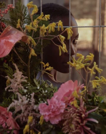 Portrait of a Black woman framed behind a wire fence and lush arrangement of pink and yellow flowers by Andreas Ortner