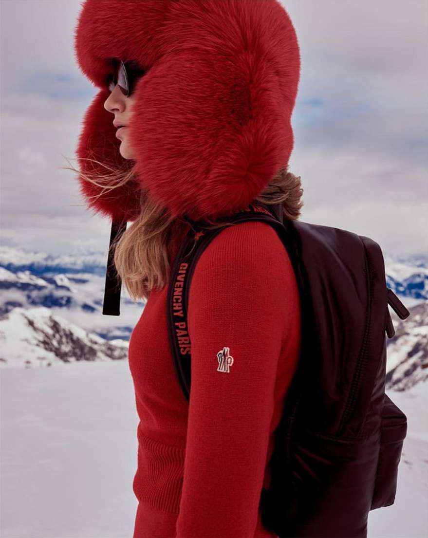 Fashionable woman in red sweater and fur hat stands in a snowy mountain landscape with a Givenchy backpack, exuding alpine style.