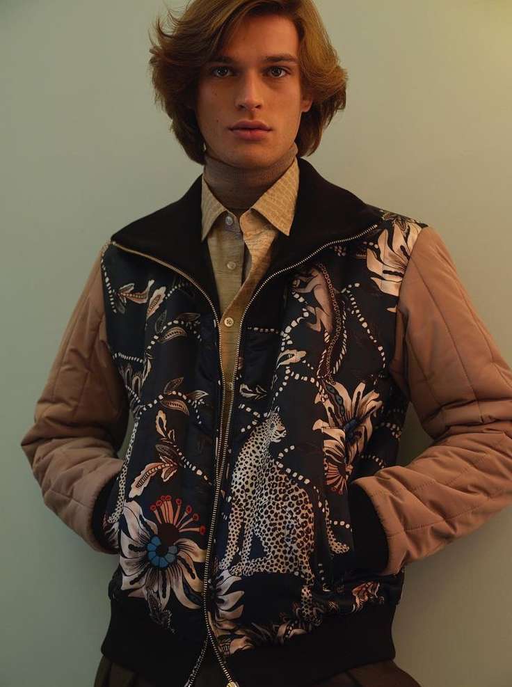 Fashionable adult male model with long brown hair wears a patterned bomber jacket, captured in a studio setting with soft, diffused lighting.