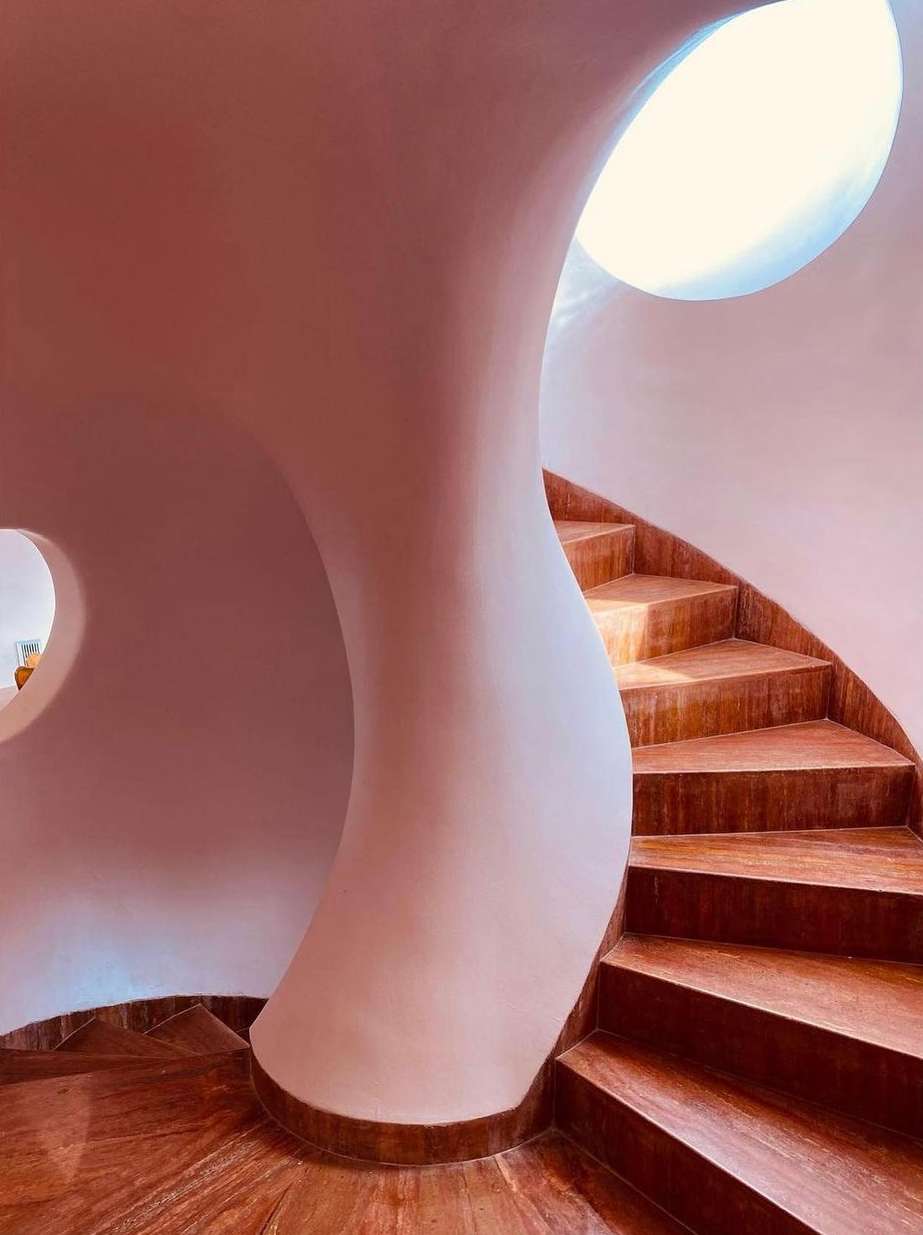 Interior view of a curving staircase with light streaming from a round skylight, set against a textured, light pink wall.
