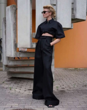 Fashionable woman poses in black crop top and wide-leg pants near a concrete staircase with an orange wall, showcasing a modern, edgy look.
