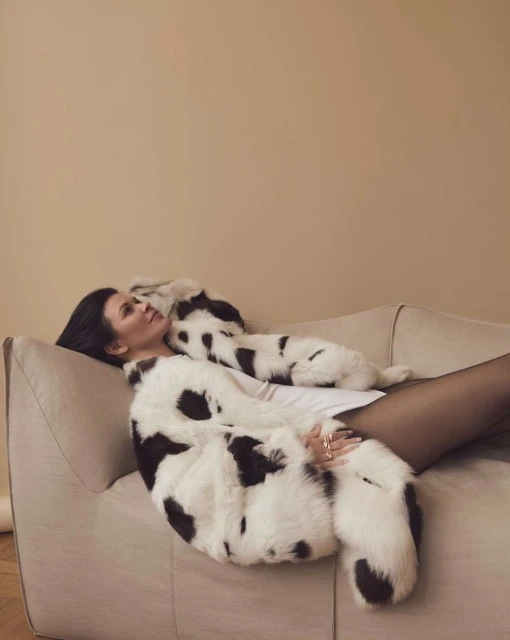 An adult female lies on a couch wearing a white and black spotted fur coat, black tights, and rings on her hand in a photo studio.