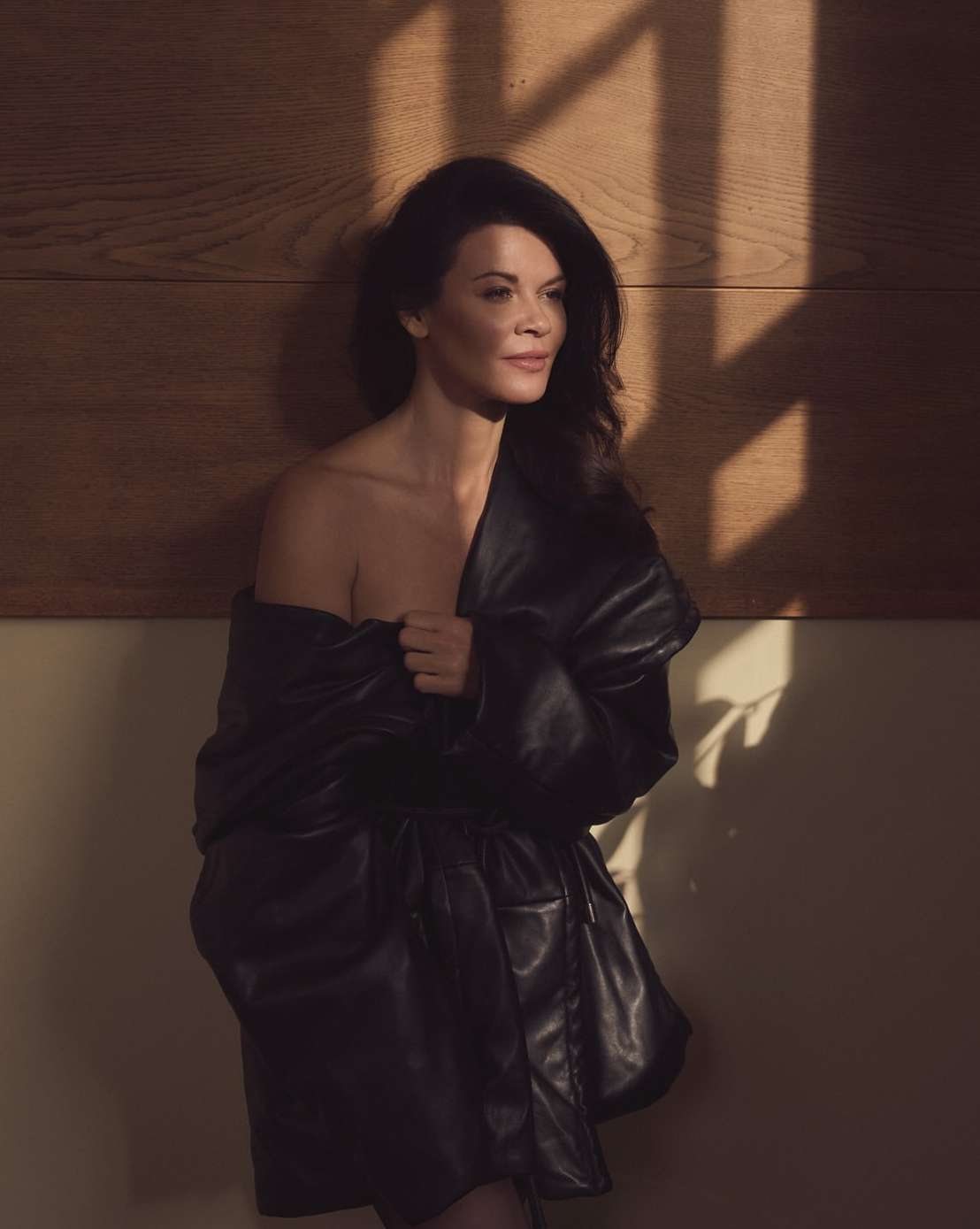 Portrait of a woman with long dark hair posing in a black leather jacket in a dimly lit indoor studio against a wood-paneled wall.