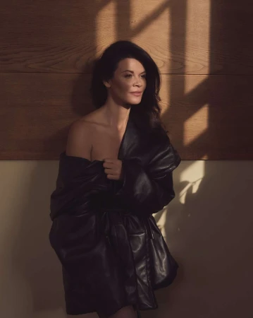 Portrait of a woman with long dark hair posing in a black leather jacket in a dimly lit indoor studio against a wood-paneled wall.