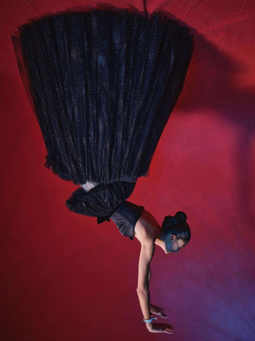 A woman in a black tulle dress performs an acrobatic handstand against a red backdrop with dramatic lighting, showcasing a blend of fashion and fitness.