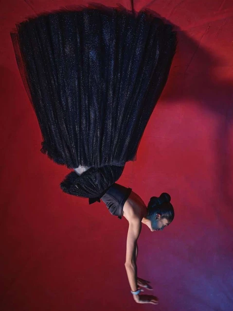 A woman in a black tulle dress performs an acrobatic handstand against a red backdrop with dramatic lighting, showcasing a blend of fashion and fitness.